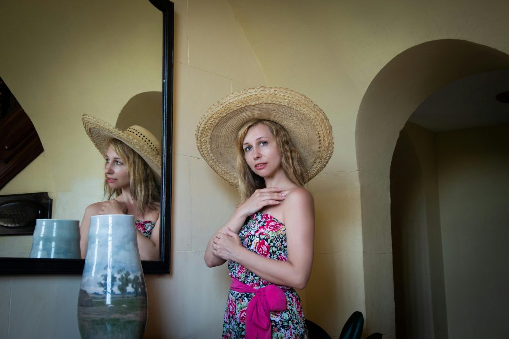 Young woman in a vintage floral dress and straw hat posing indoors by a mirror.