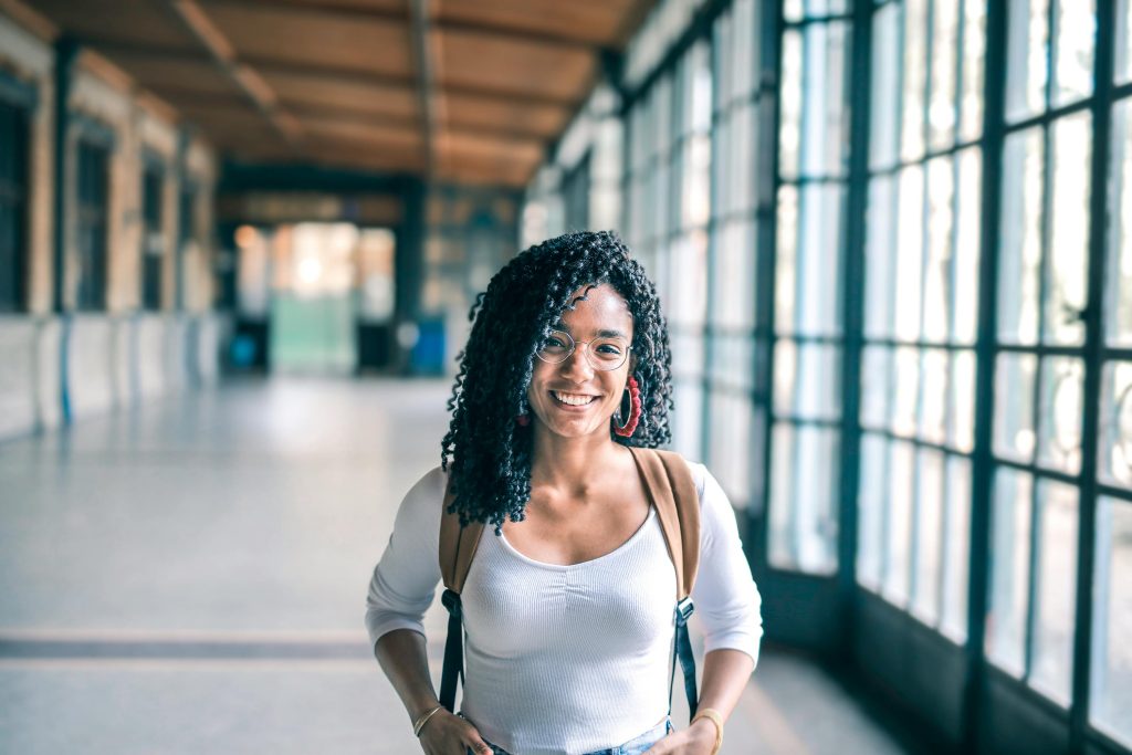 A cheerful woman with glasses stands in a bright indoor hallway, carrying a backpack.